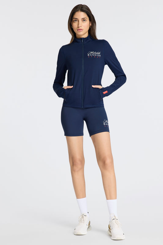 Model standing with hands in her pockets wearing Bandier x Michelob ULTRA Full Zip Jacket, "Michelob BANDIER Sport" on left chest, Michelob ULTRA ribbon on left sleeve near wrist