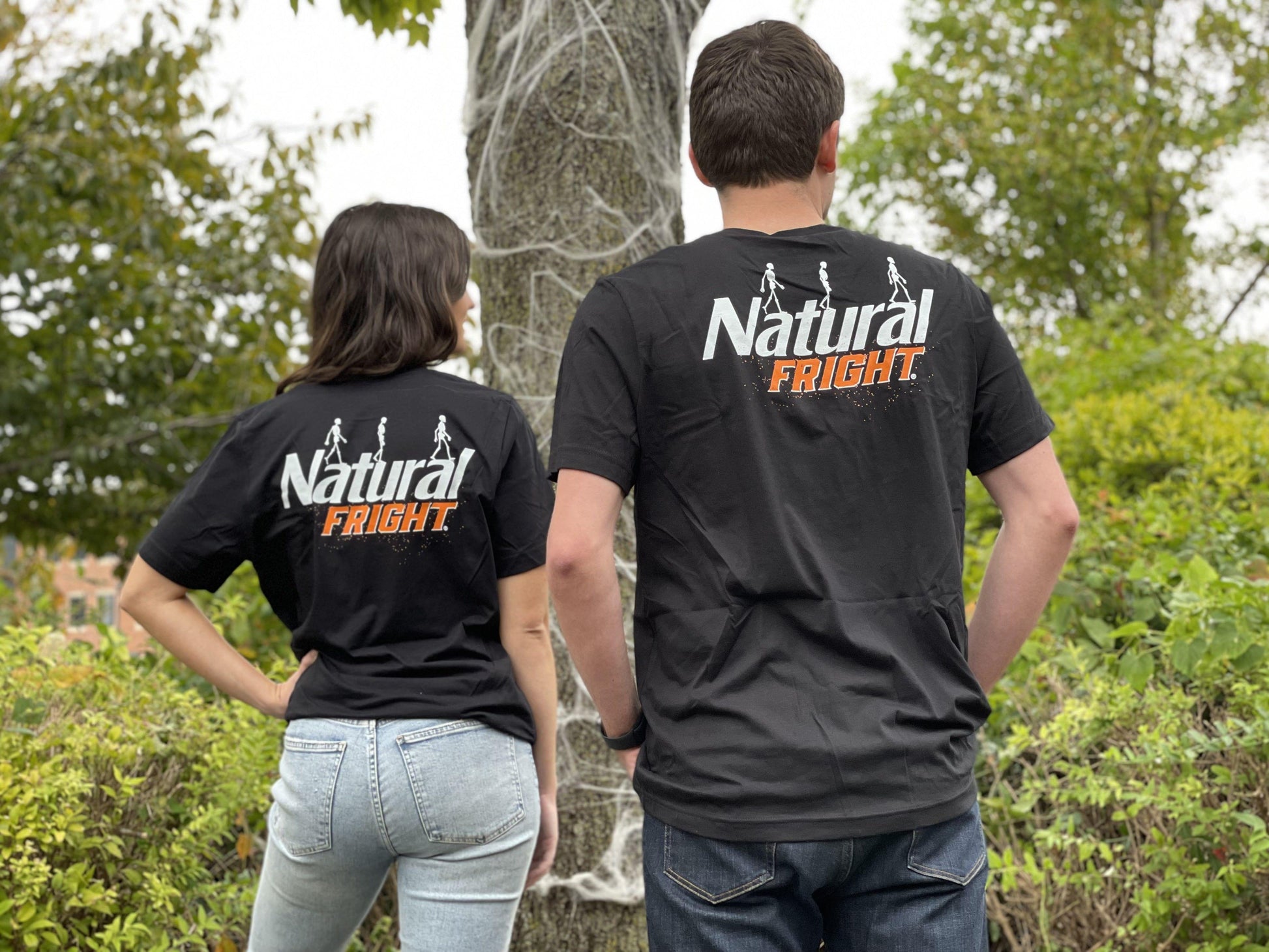woman and man wearing black natural light backprint t shirt with natural light logo in white and orange on the back of shirt with white skeletons walking across logo
