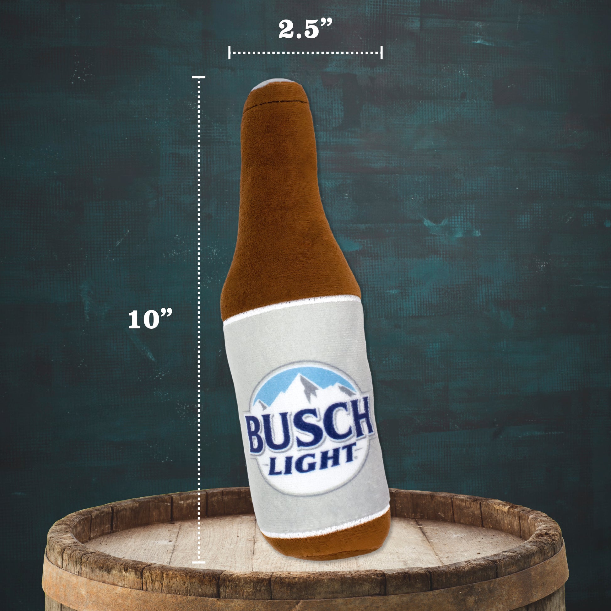 A plush beer bottle toy styled like a Busch Light bottle, shown leaning on a wooden barrel with size annotations indicating 10 inches tall and 2.5 inches wide, against a dark textured background.
