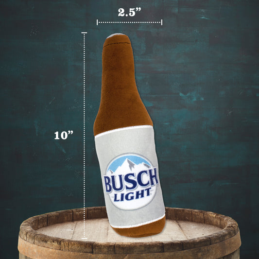 A plush beer bottle toy styled like a Busch Light bottle, shown leaning on a wooden barrel with size annotations indicating 10 inches tall and 2.5 inches wide, against a dark textured background.