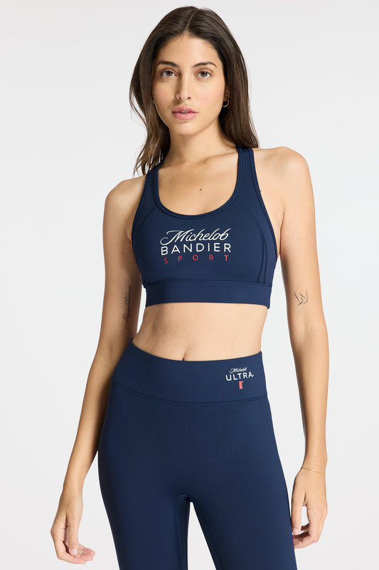 Front view of a model wearing Bandier x Michelob Ultra Center Stage Bra, text "Michelob Bandier Sport" across the chest, and wearing Michelob x Bandier Leggings