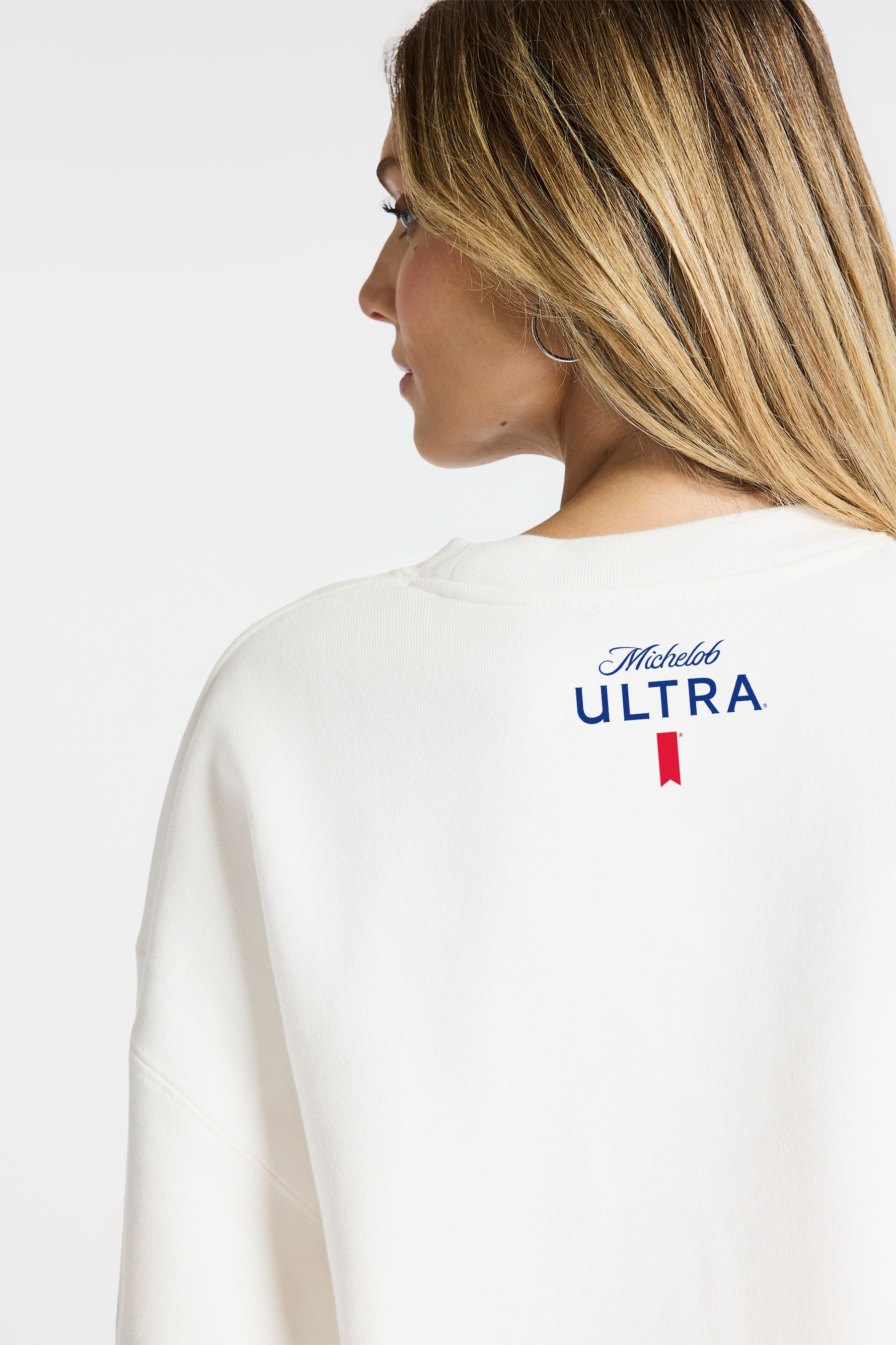 Back view of Model wearing Bandier x Michelob ULTRA Sweatshirt, Michelob ULTRA logo and ribbon at top of center back sweatshirt