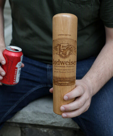 A person sitting on stone steps holds a Budweiser-branded wooden cylinder-shaped object in one hand while holding a red beverage can in the other.